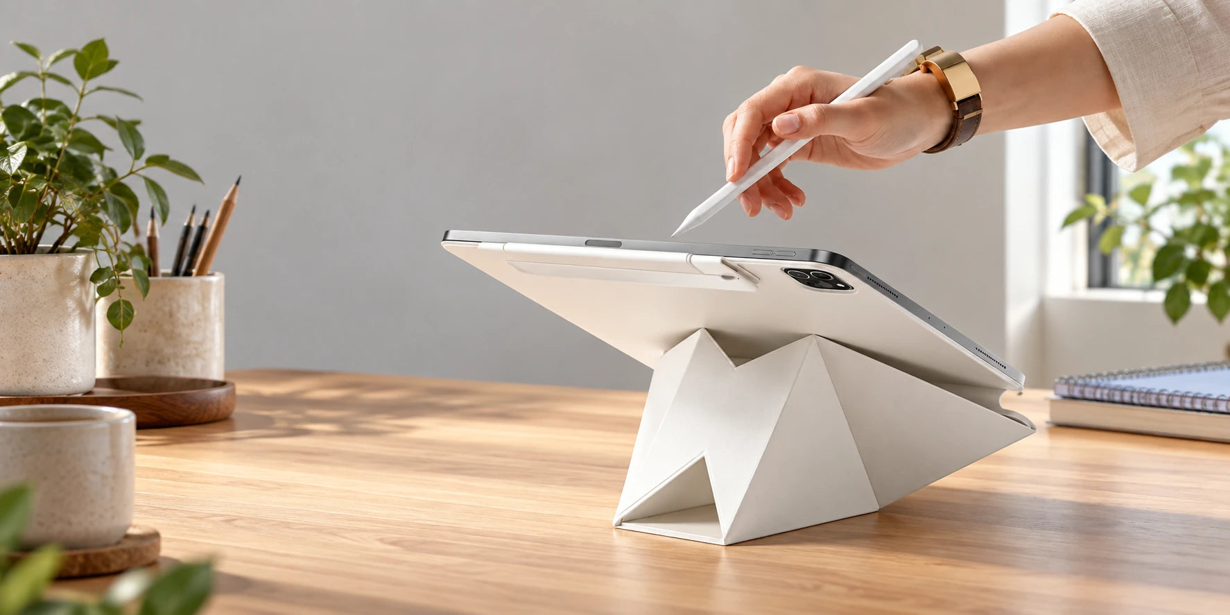 Hand using a white stylus with a tablet supported at a low angle by an origami stand.