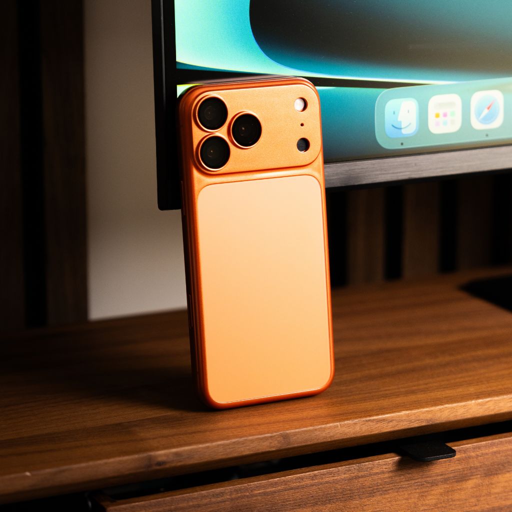 A matte orange phone case shown from the back, standing upright on a wooden desk with a computer monitor in the blurred background.